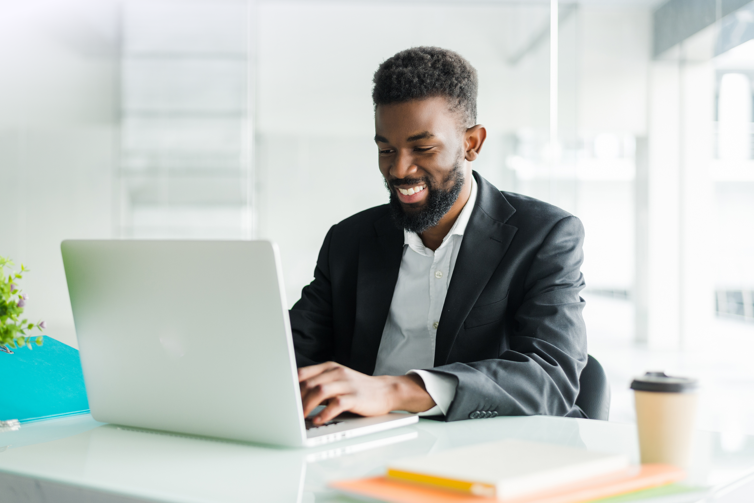 A man sitting at a desk in a bright modern office, working on a laptop with notebooks and a coffee cup on the table.